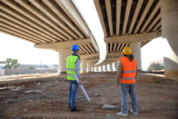 Two men wearing hard hats and high vis stood under a bridge structure with one man holding a roll of paper