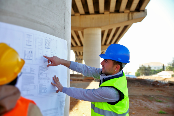 A man in a blue hard hat pointing at infrastructure plans whilst another person looks on
